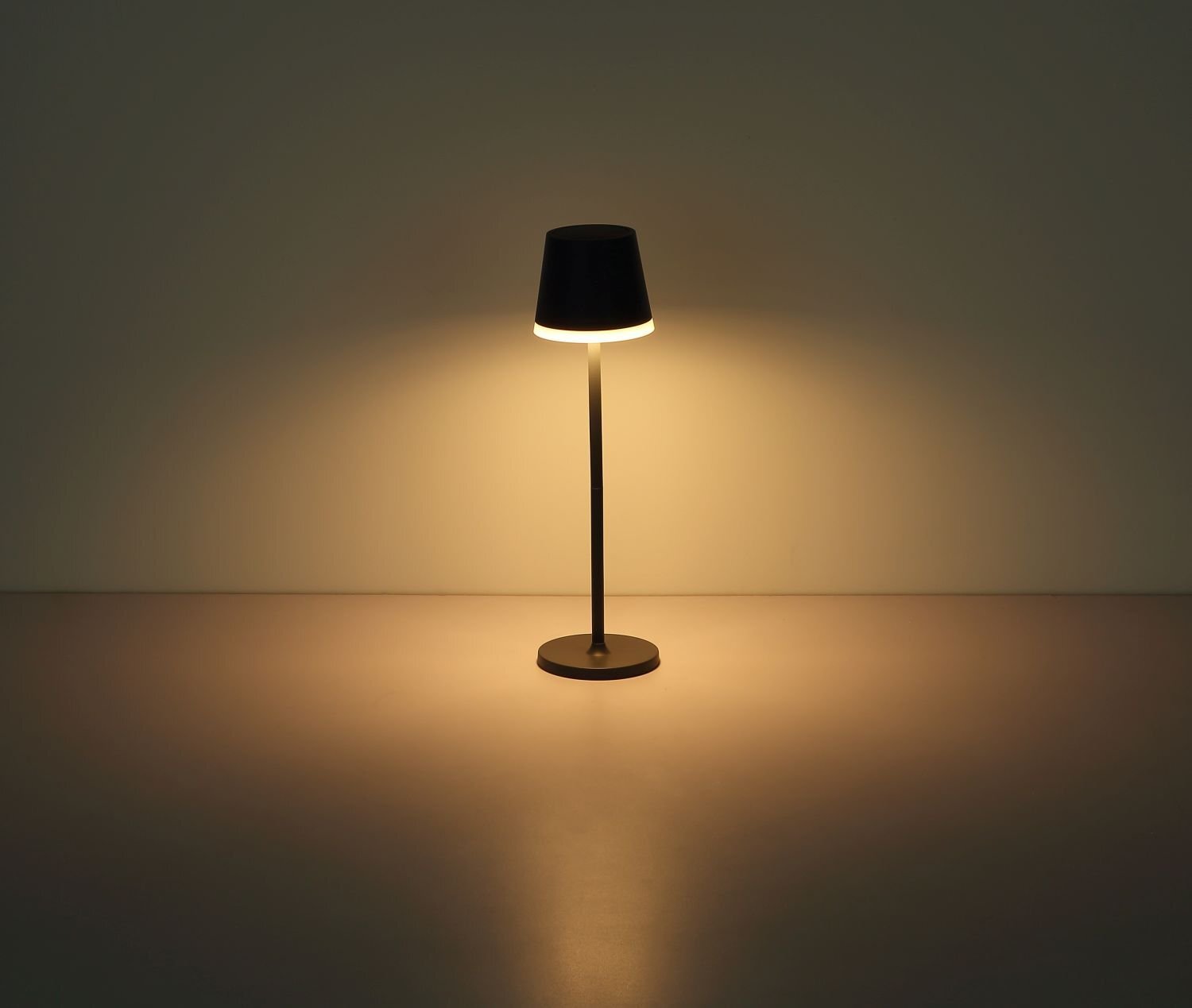 Na obrazie widoczna jest włączona lampa stołowa z ciemnym kloszem, emitująca ciepłe światło na powierzchnię poniżej. Lampa stoi na gładkim, jednolitym tle.