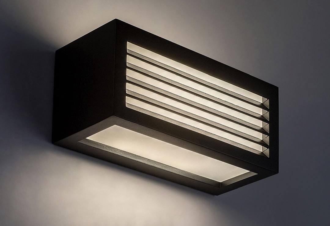 Czarna, prostokątna lampa ścienna z poziomymi szczelinami, emitująca białe światło. Lampa jest zamontowana na ścianie i oświetla przestrzeń wokół siebie.