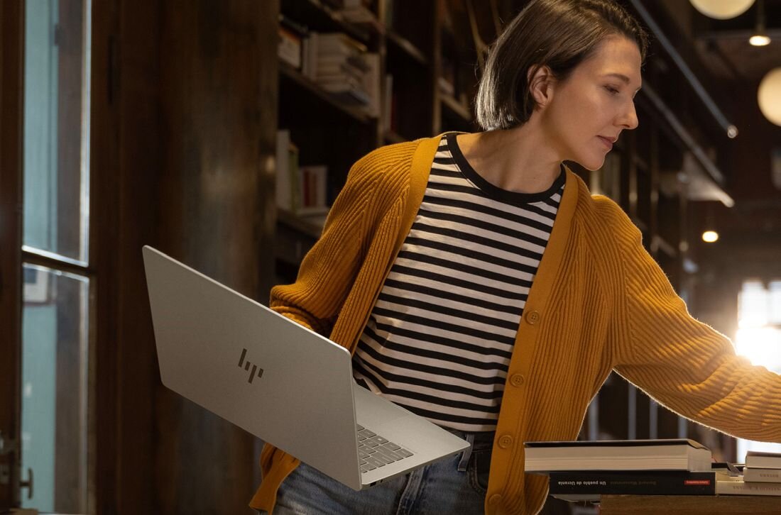 Osoba trzymająca srebrny laptop z widocznym logo HP, ubrana w żółty sweter i koszulkę w paski, stoi w pomieszczeniu z półkami pełnymi książek.