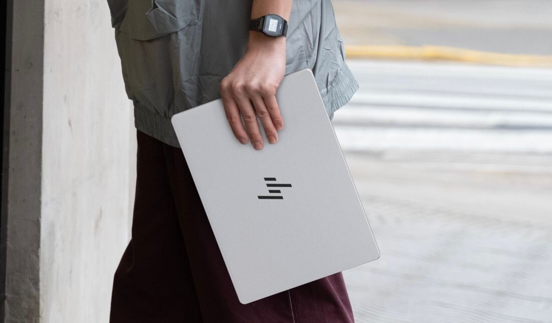 Osoba trzyma biały laptop z widocznym logo w formie poziomych linii. W tle widać fragment chodnika i ulicy.
