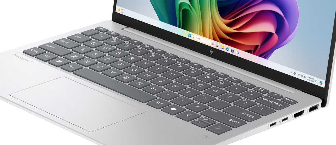 Na obrazie widoczny jest srebrny laptop z otwartym ekranem, na którym wyświetlany jest kolorowy motyw. Klawiatura ma ciemne klawisze z białymi oznaczeniami, a na obudowie znajduje się logo producenta.