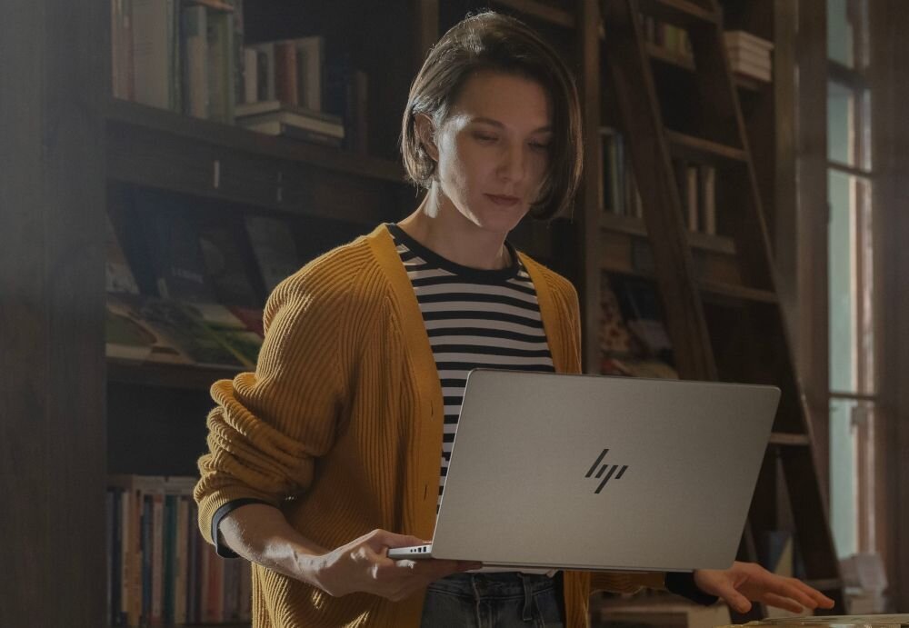 Osoba trzymająca laptopa z widocznym logo HP, w tle regał z książkami.