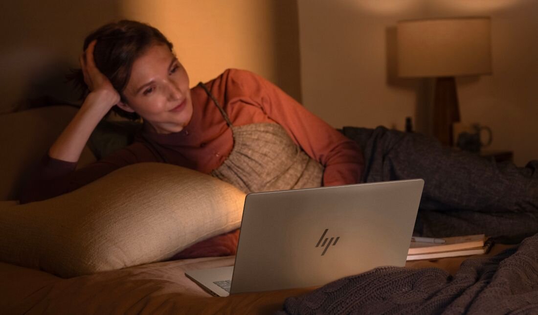 Osoba leży na łóżku, opierając się na poduszce, i patrzy na otwarty laptop z widocznym logo HP. W tle znajduje się lampa stołowa.
