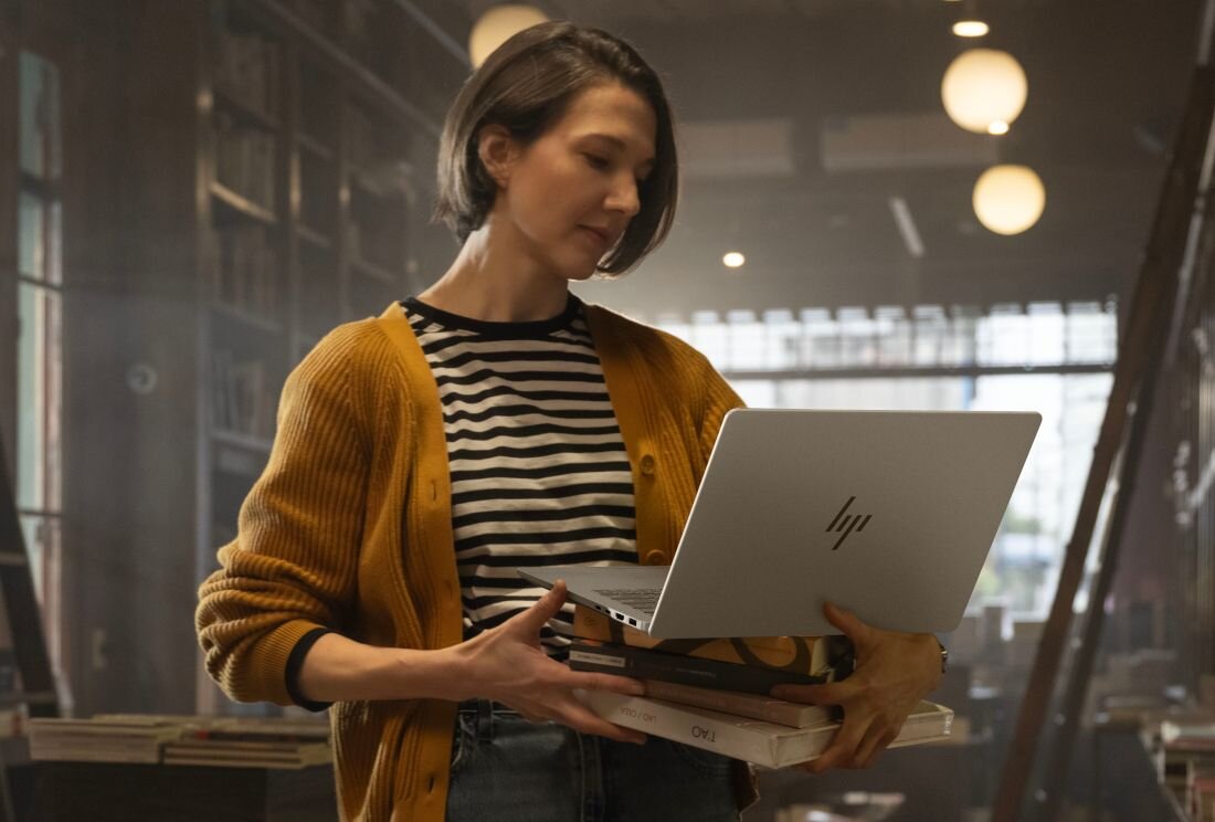 Osoba trzymająca otwarty laptop z widocznym logo HP, stojąca w pomieszczeniu przypominającym bibliotekę lub księgarnię. W tle widać półki z książkami i okrągłe lampy sufitowe.