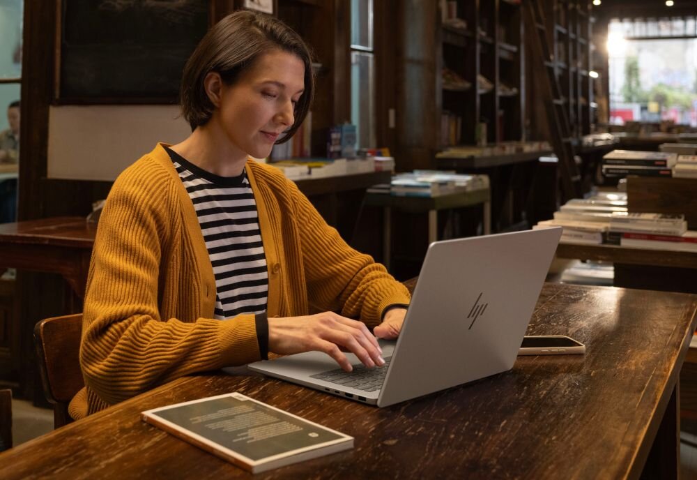 Osoba siedzi przy drewnianym stole, używając laptopa z logo HP. W tle widać półki z książkami i inne meble.