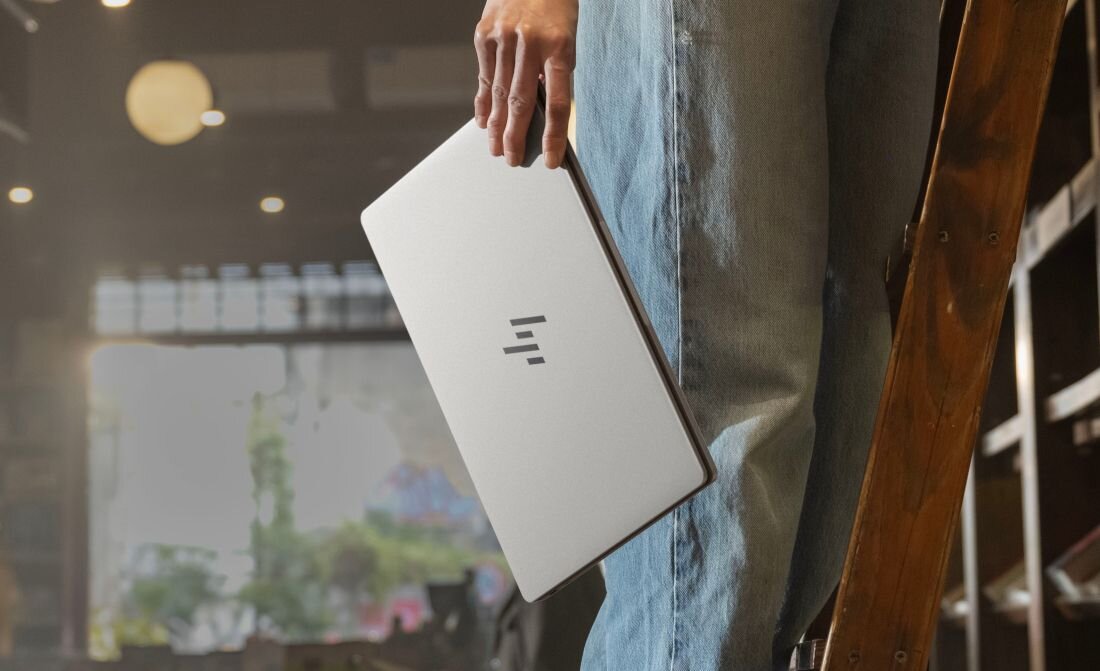 Osoba trzyma zamknięty laptop z widocznym logo producenta. W tle znajduje się wnętrze sklepu media expert.