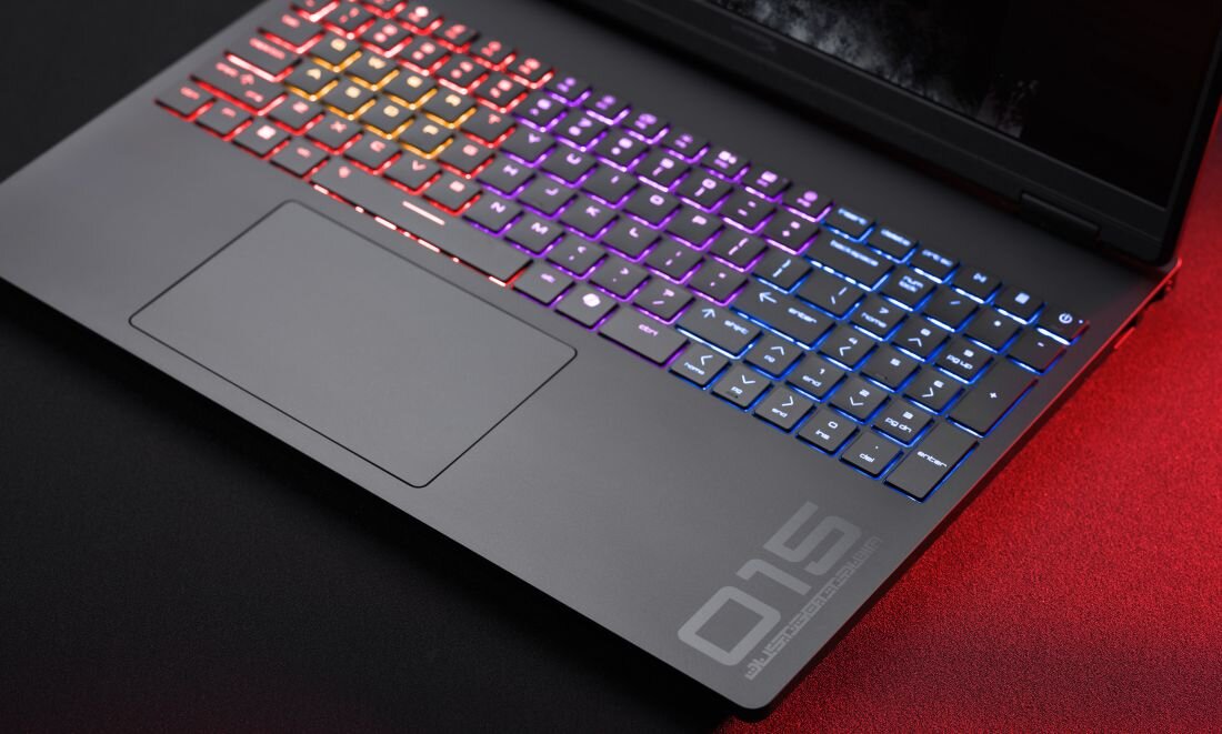 Laptop z podświetlaną klawiaturą w różnych kolorach, z widocznym napisem \'015\' na obudowie. Powierzchnia wokół touchpada jest matowa, a tło jest ciemne z czerwonym oświetleniem.
