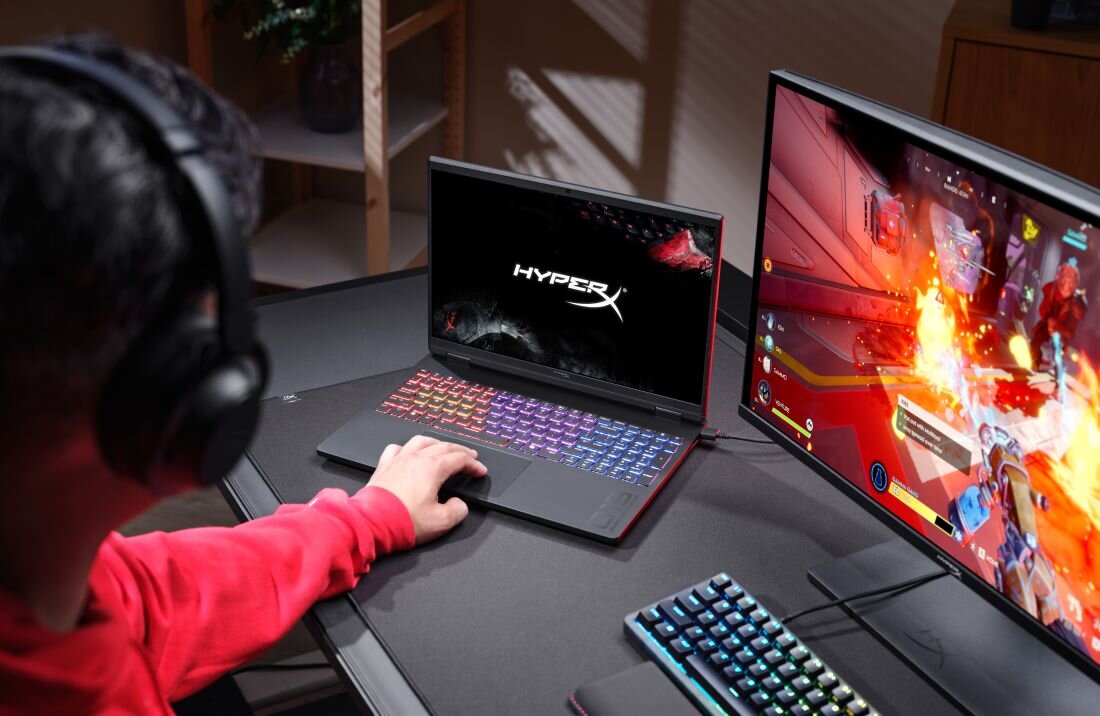 Osoba siedzi przy biurku, używając laptopa z podświetlaną klawiaturą, na ekranie którego widnieje logo \'HyperX\'. Obok znajduje się monitor z wyświetloną grą komputerową.