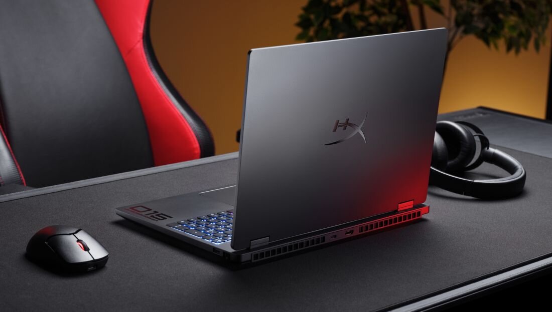 Na biurku znajduje się otwarty laptop z logo \'HX\' na pokrywie, obok myszka i słuchawki. W tle widoczny jest fragment czerwono-czarnego fotela oraz rośliny.