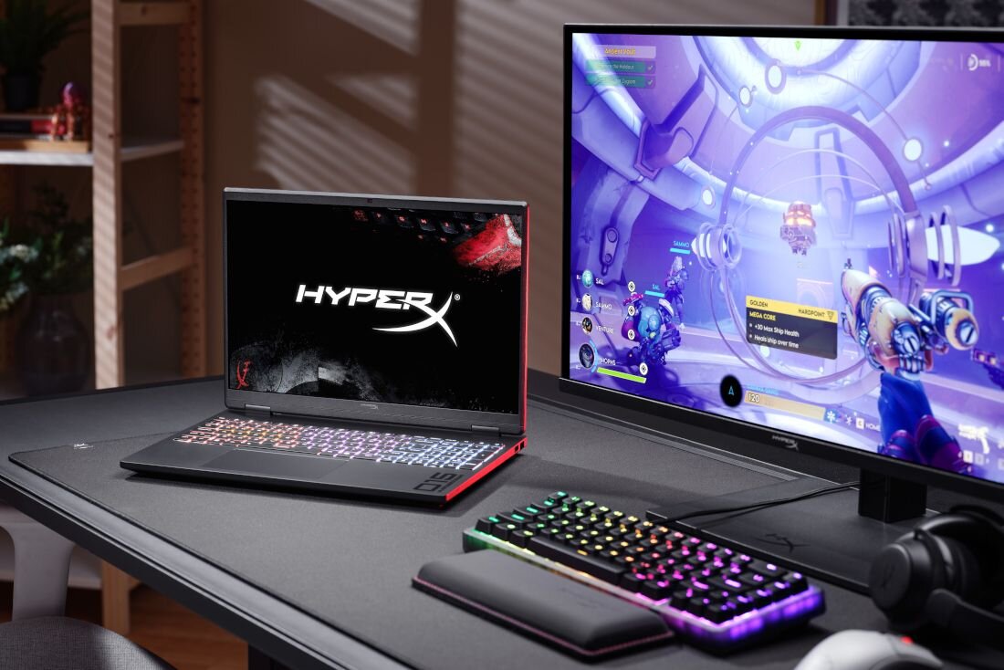 Na biurku znajduje się laptop z logo \'HyperX\' na ekranie oraz monitor wyświetlający grę. Obok laptopa leży klawiatura i myszka z podświetleniem RGB.