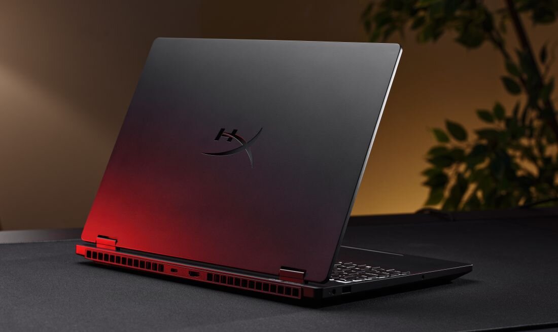 Laptop z logo \'HX\' na pokrywie, ustawiony na ciemnym stole z czerwonym podświetleniem. W tle widoczne są rozmyte rośliny.
