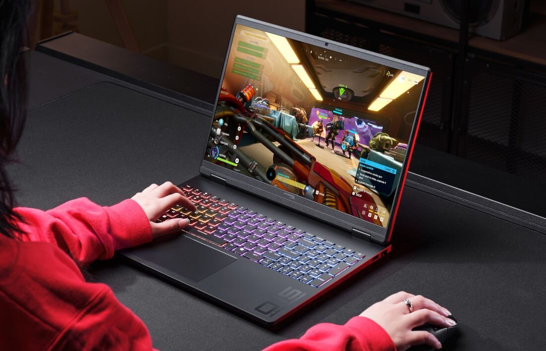 Osoba używa laptopa z kolorową klawiaturą, na ekranie widoczna jest gra komputerowa. Laptop znajduje się na ciemnym biurku.
