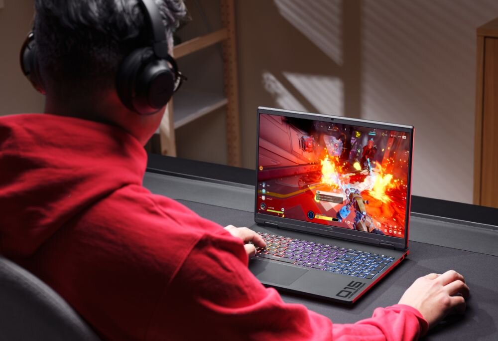 Osoba w czerwonej bluzie i słuchawkach gra na laptopie z kolorową klawiaturą, na ekranie widoczna jest dynamiczna gra akcji. Na obudowie laptopa znajduje się logo \'Omen\'.