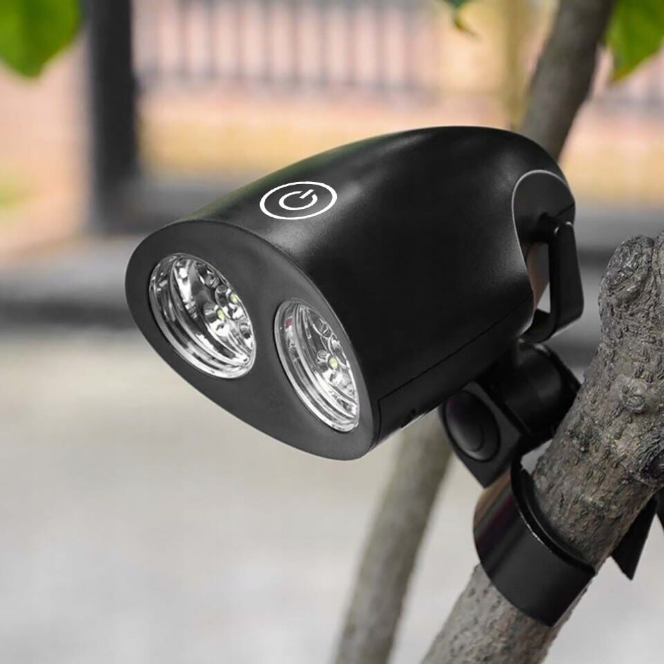 Czarna lampa LED z dwoma reflektorami zamontowana na gałęzi drzewa. Na górze lampy widoczny jest symbol włącznika.