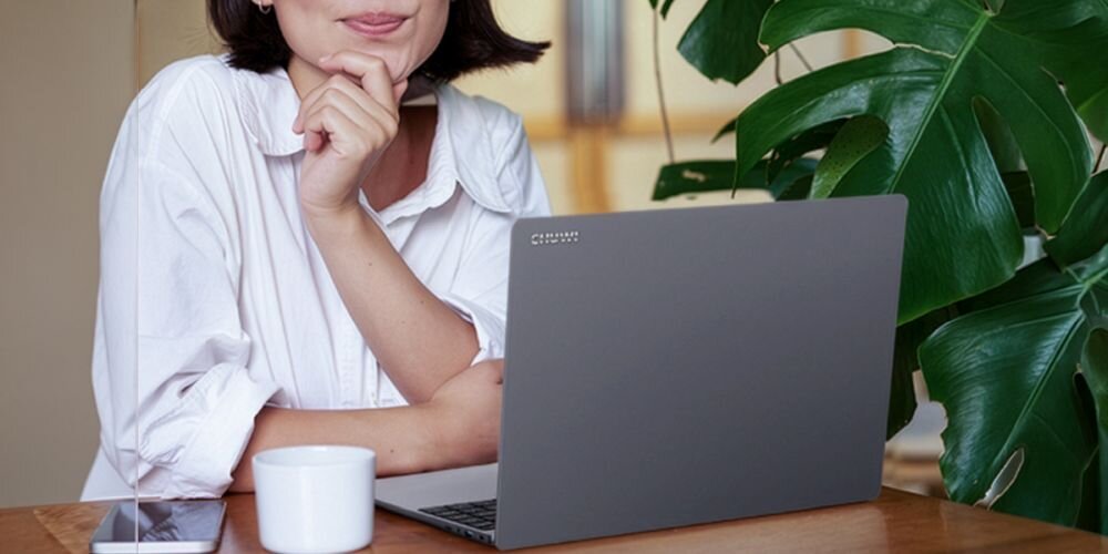 Osoba siedzi przy laptopie na stole obok kubka telefonu i dużej zielonej rośliny