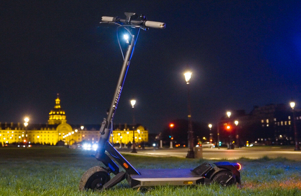Czarna hulajnoga elektryczna stoi na trawie w nocy, oświetlona latarniami ulicznymi. W tle widoczny jest oświetlony budynek z kopułą.