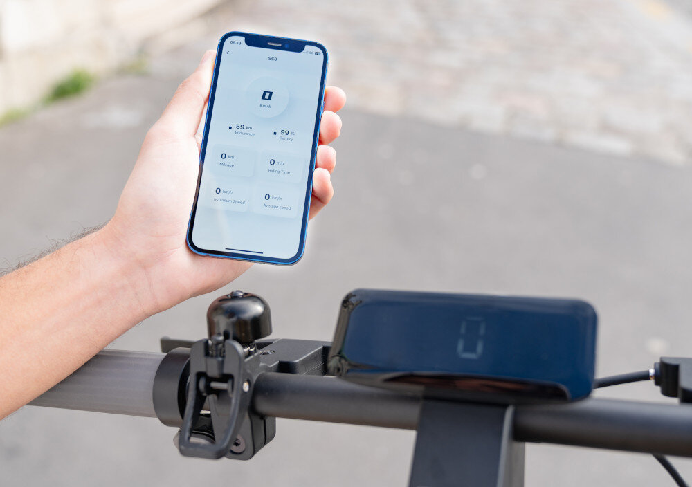 Osoba trzyma smartfon z aplikacją pokazującą dane dotyczące hulajnogi elektrycznej. Na kierownicy hulajnogi znajduje się wyświetlacz z cyfrą 0.