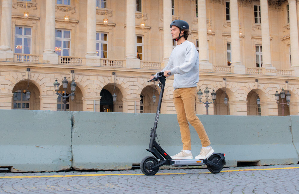 Osoba w kasku jedzie na czarnej hulajnodze elektrycznej przed budynkiem z kolumnami. Hulajnoga ma duże koła i widoczny napis na kierownicy.
