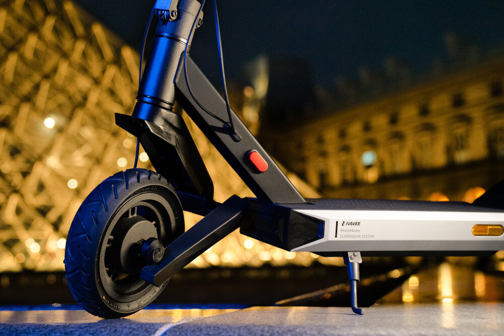 Zbliżenie na przednie koło czarnej hulajnogi elektrycznej z widocznym napisem \'NAVEE Suspension System\'. W tle rozmyte światła budynku.