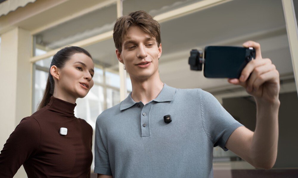 Dwie osoby stoją obok siebie, każda z nich ma przypięty mały, kwadratowy nadajnik na ubraniu. Jedna z osób trzyma smartfon z zamontowanym mikrofonem, robiąc zdjęcie lub nagrywając film.