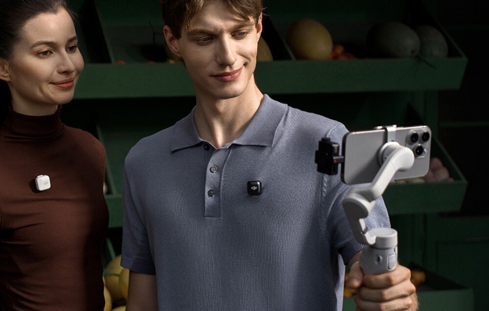 Mężczyzna trzyma smartfon zamocowany na stabilizatorze, a na jego koszulce przypięty jest czarny nadajnik z logo DJI. Obok stoi kobieta z białym urządzeniem przypiętym do ubrania.