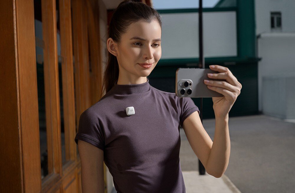 Osoba trzymająca smartfon robi zdjęcie, mając przypięty mały, czarny nadajnik na koszulce. W tle widoczne są drewniane drzwi i fragment budynku.