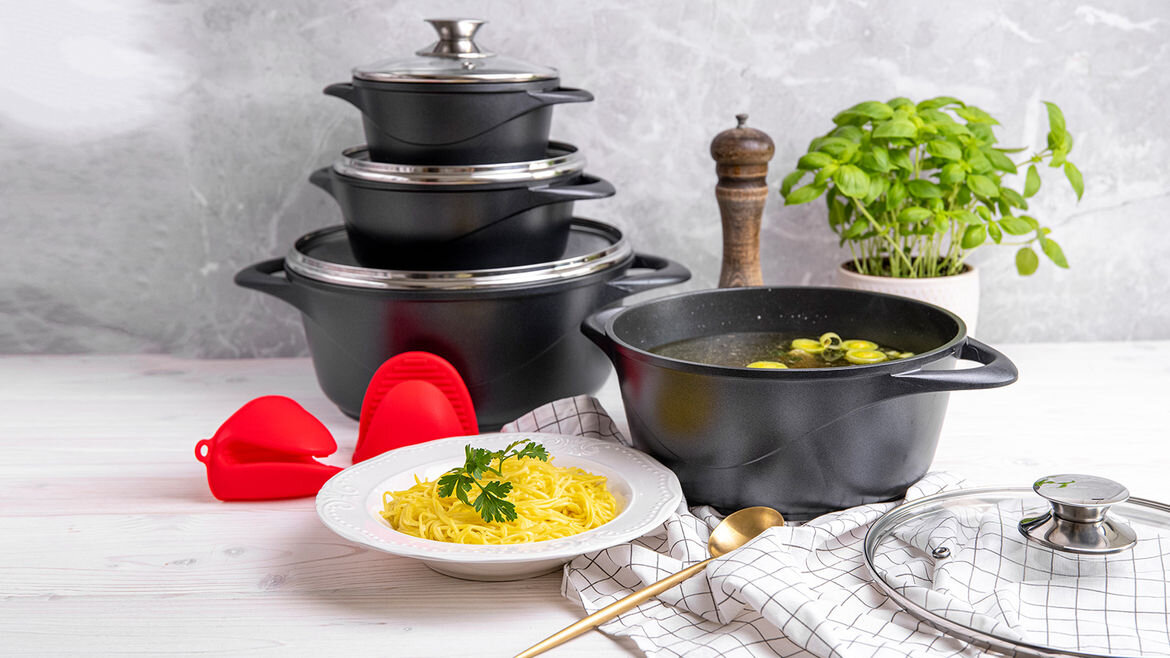 Na obrazie widoczny jest zestaw czarnych garnków z pokrywkami, ustawionych jeden na drugim, obok talerza z makaronem i ziołami. W tle znajduje się doniczka z bazylią i młynek do pieprzu.