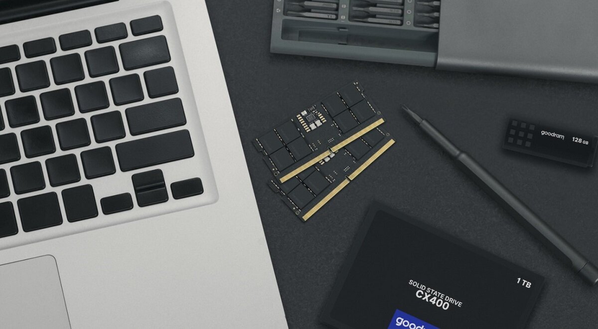 Na ciemnym tle znajduje się moduł pamięci RAM z widocznymi chipami, obok leży laptop, długopis oraz dysk SSD z napisem \'SOLID STATE DRIVE CX400 1 TB\'.