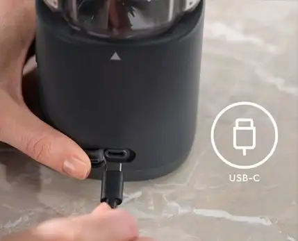 Osoba podłącza kabel USB-C do czarnego urządzenia stojącego na marmurowym blacie. Obok znajduje się symbol USB-C z napisem \'USB-C\'.