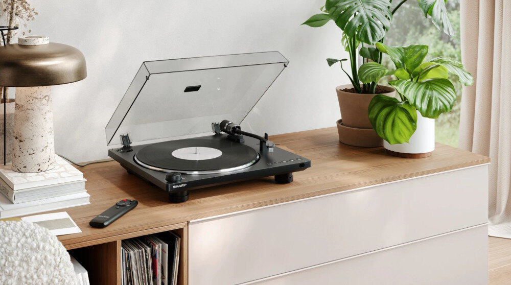Na drewnianej komodzie stoi czarny gramofon z przezroczystą pokrywą, obok którego leży pilot. W tle znajdują się rośliny doniczkowe i lampa.