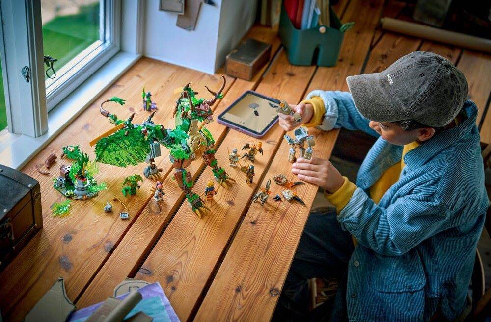 Na drewnianym stole znajduje się zestaw klocków LEGO przedstawiający zielonego smoka i kilka figurek. Obok zestawu siedzi dziecko w czapce, bawiące się klockami.