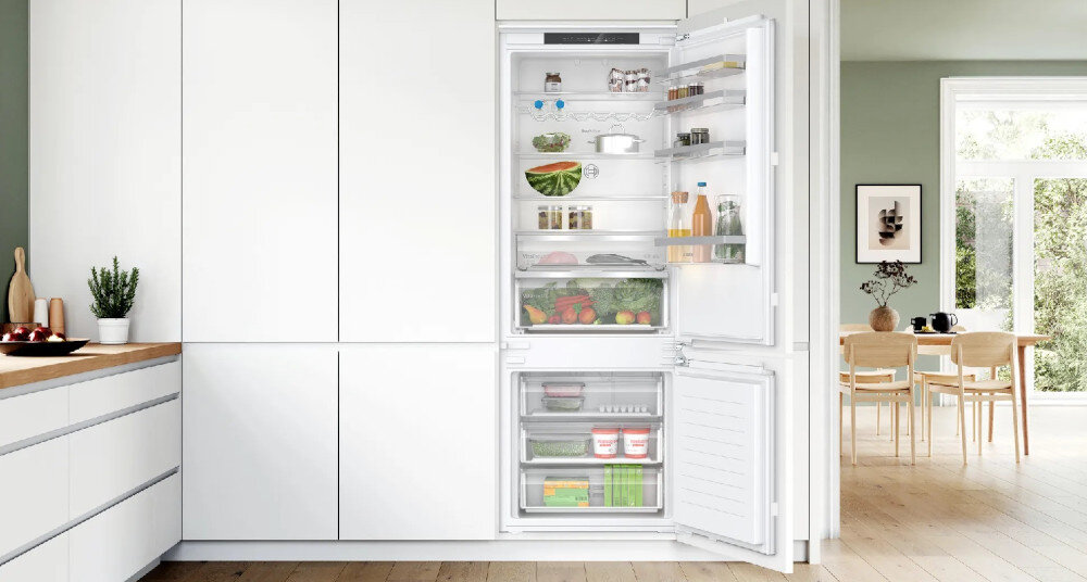Wnętrze nowoczesnej kuchni z otwartą lodówką w zabudowie, wypełnioną różnorodnymi produktami spożywczymi. W tle widoczny jest stół z krzesłami oraz okno z widokiem na zewnątrz.