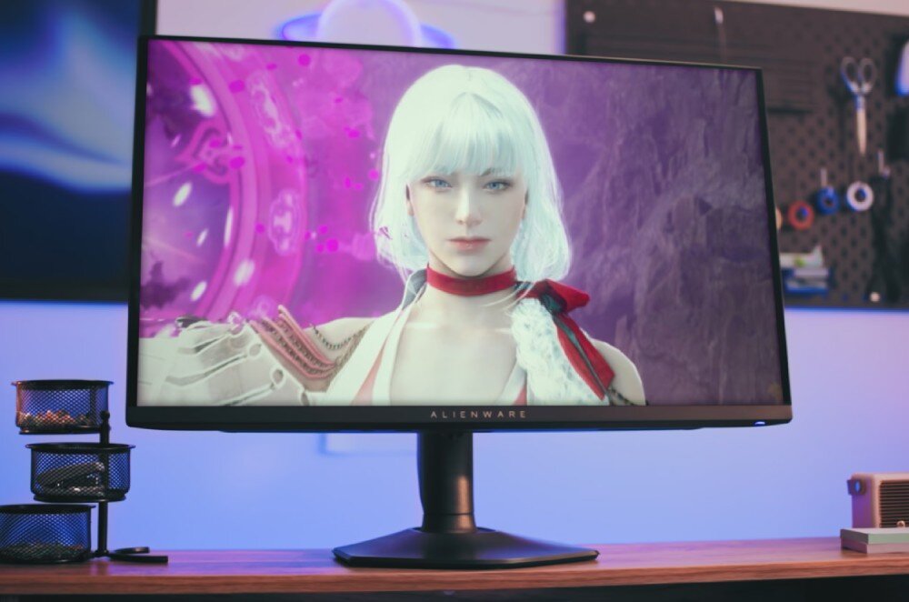 Monitor marki Alienware wyświetla postać z białymi włosami na tle fioletowych elementów graficznych. Na dolnej ramce monitora widoczne jest logo \'ALIENWARE\'.