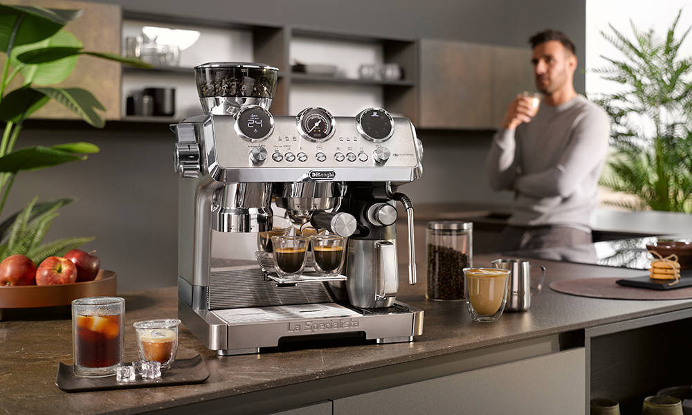 Na blacie kuchennym stoi ekspres do kawy z logo \'DeLonghi\', obok którego znajdują się filiżanki z kawą i szklanki z napojami. W tle widać osobę trzymającą filiżankę.