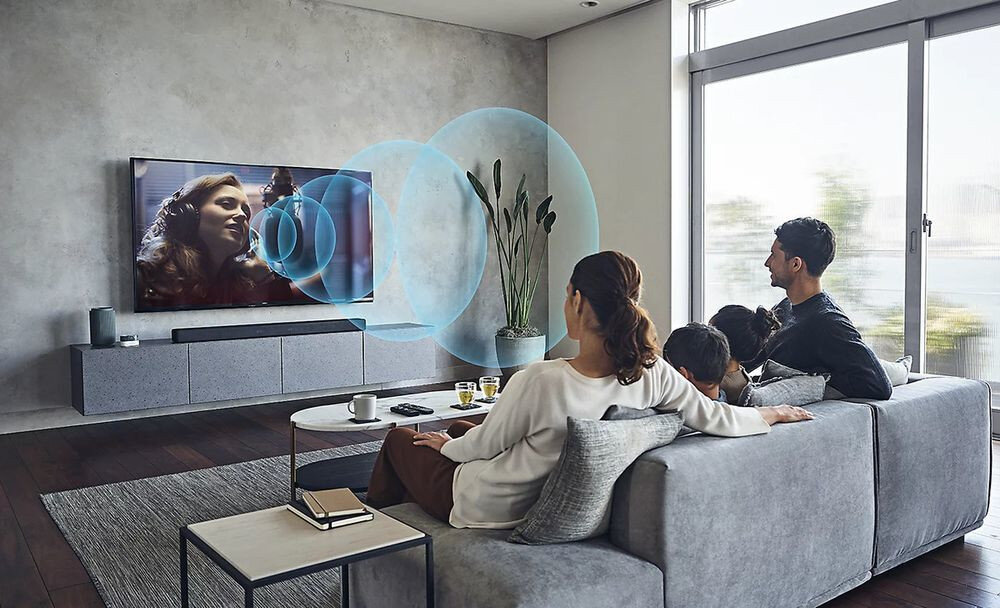 W salonie na ścianie wisi duży telewizor, na którym wyświetlany jest obraz kobiety w słuchawkach. Przed telewizorem siedzi rodzina na szarej kanapie, a wokół telewizora widoczne są graficzne fale dźwiękowe.