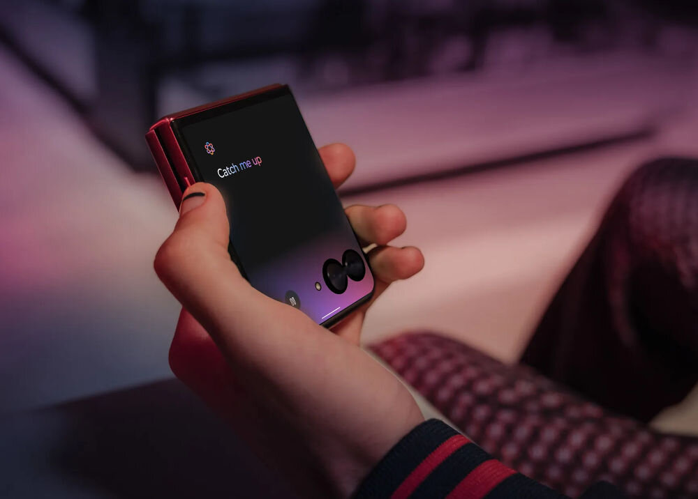 Osoba trzyma w dłoni składany smartfon z widocznym ekranem, na którym wyświetla się tekst \'Catch me up\'.