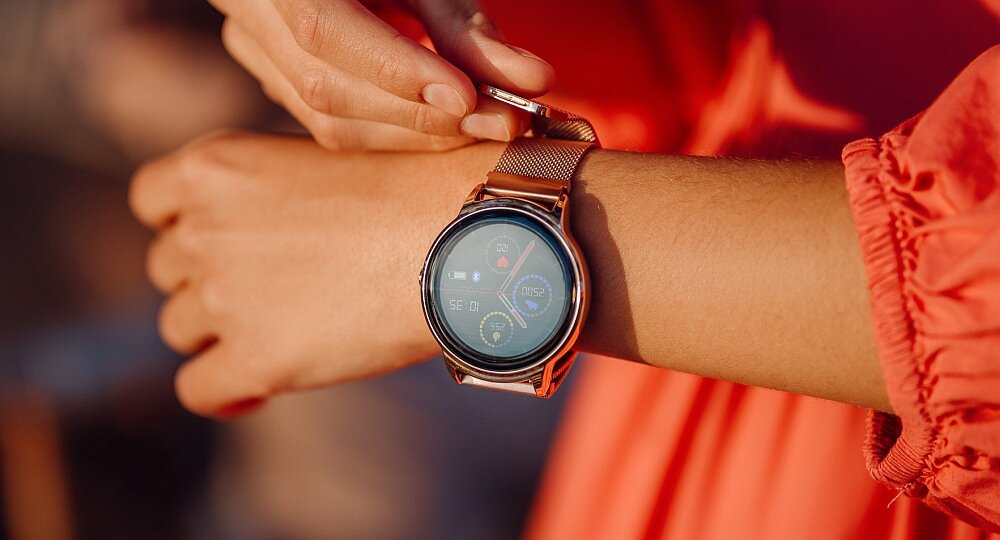 Osoba w czerwonym ubraniu zakłada smartwatch z okrągłym wyświetlaczem i metalową bransoletą. Na ekranie widoczne są kolorowe ikony i wskaźniki.