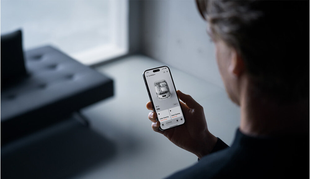 Osoba trzyma smartfon, na którym wyświetlana jest aplikacja z obrazem robota sprzątającego. W tle znajduje się rozmyta kanapa.