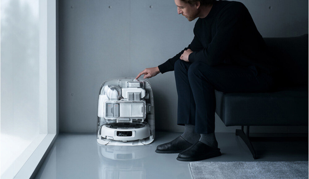 Osoba siedzi na sofie i dotyka przezroczystej obudowy, w której znajduje się biały robot sprzątający. Pomieszczenie jest minimalistycznie urządzone z szarymi ścianami i podłogą.