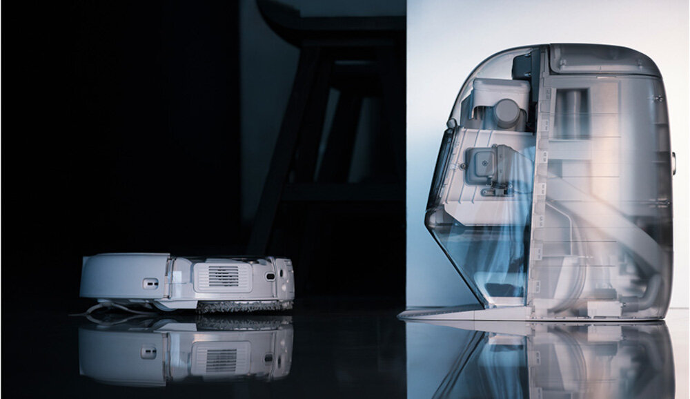 Na obrazie widoczny jest robot sprzątający oraz jego stacja dokująca z przezroczystą obudową, ukazującą wewnętrzne mechanizmy. Tło jest ciemne, a powierzchnia podłogi odbija światło.