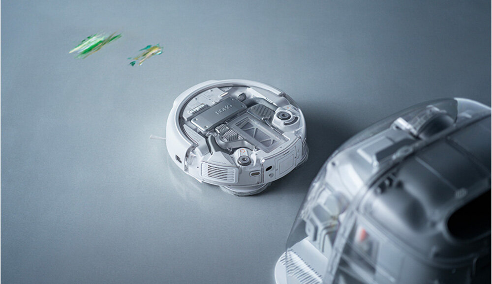 Na obrazie widoczny jest robot sprzątający o nowoczesnym designie, z przezroczystą obudową ukazującą wewnętrzne komponenty. W tle znajdują się rozmyte, kolorowe elementy.