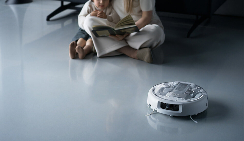 Na podłodze znajduje się biały robot sprzątający. W tle siedzą dwie osoby, jedna z nich trzyma otwartą książkę.