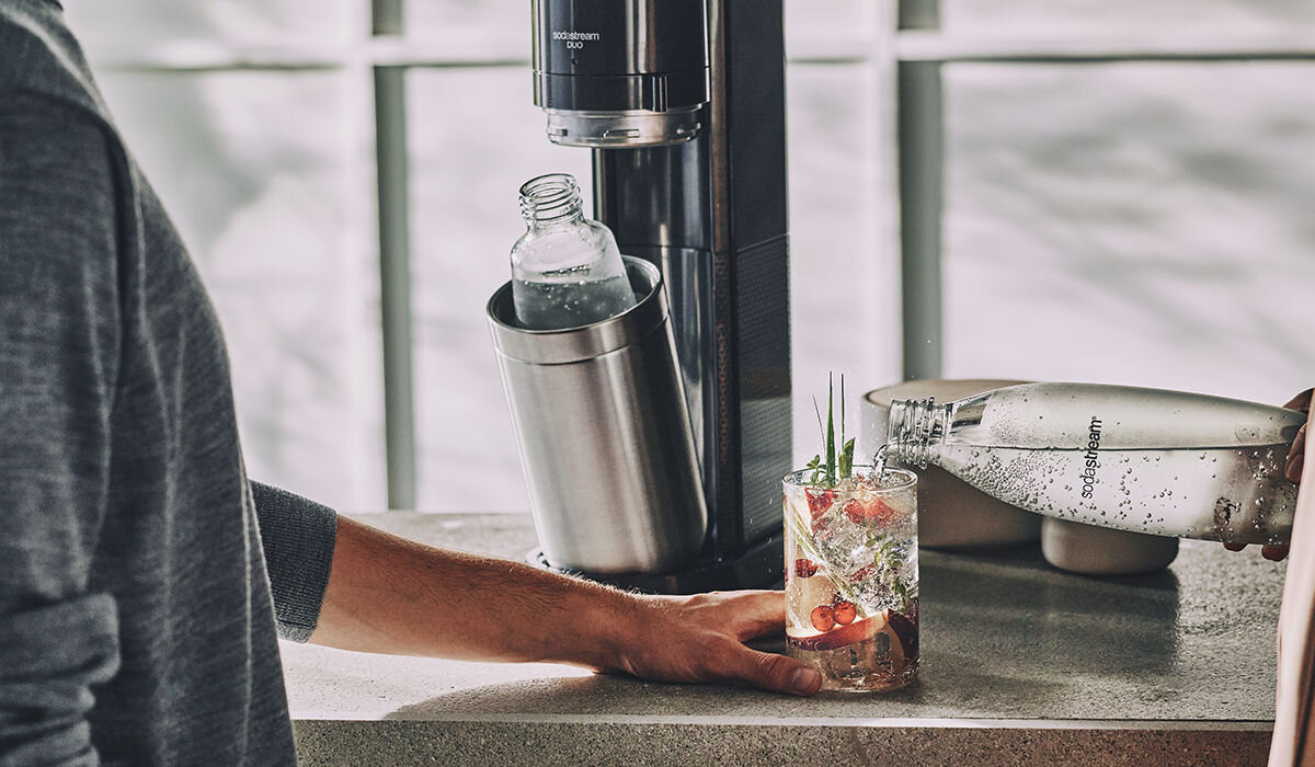 Na blacie kuchennym stoi saturator z butelką w środku, obok znajduje się szklanka z napojem i owocami. Osoba nalewa wodę z butelki z logo \'SodaStream\'.