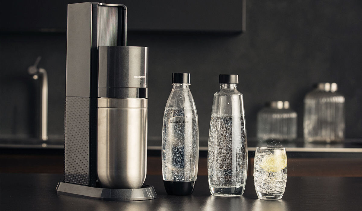Na obrazie znajduje się czarny saturator z metalicznym akcentem oraz dwie przezroczyste butelki z wodą gazowaną i szklanka z napojem. Na saturatorze widoczne jest logo \'SODASTREAM\'.