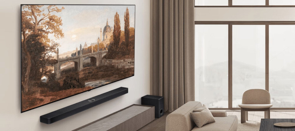 W salonie na ścianie wisi telewizor wyświetlający obraz przedstawiający most i drzewa. Pod telewizorem znajduje się soundbar, a w pomieszczeniu są także sofa i stolik.