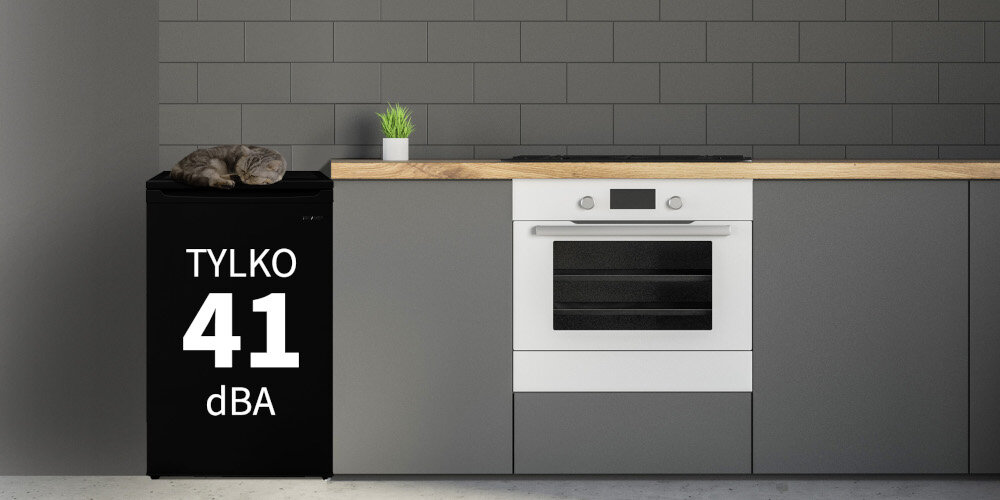 Czarna lodówka z napisem \'TYLKO 41 dBA\' stoi obok szarej zabudowy kuchennej z piekarnikiem i blatem, na którym znajduje się roślina w doniczce.