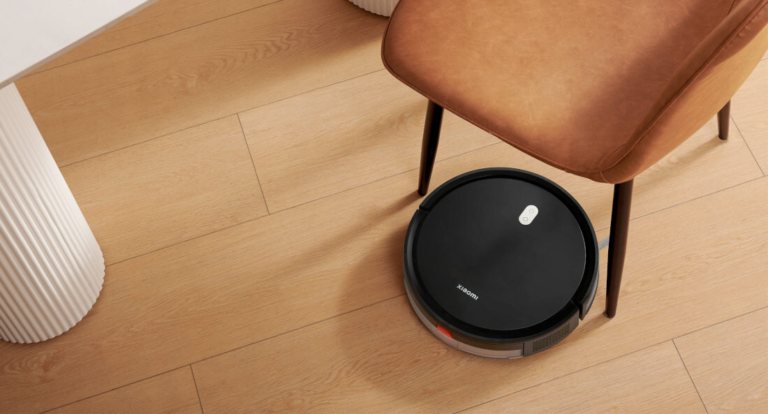 Czarny robot sprzątający znajduje się na drewnianej podłodze obok brązowego krzesła. Na robocie widoczny jest napis \'xiaomi\'.