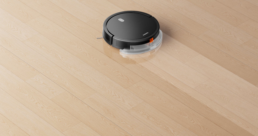 Czarny robot sprzątający porusza się po jasnej, drewnianej podłodze. Na urządzeniu widoczne są dwa przyciski i logo producenta.