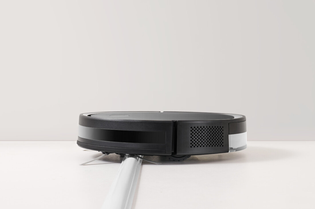 Czarny robot sprzątający porusza się po jasnej podłodze. Urządzenie ma okrągły kształt i widoczne są jego sensory oraz kratki wentylacyjne.