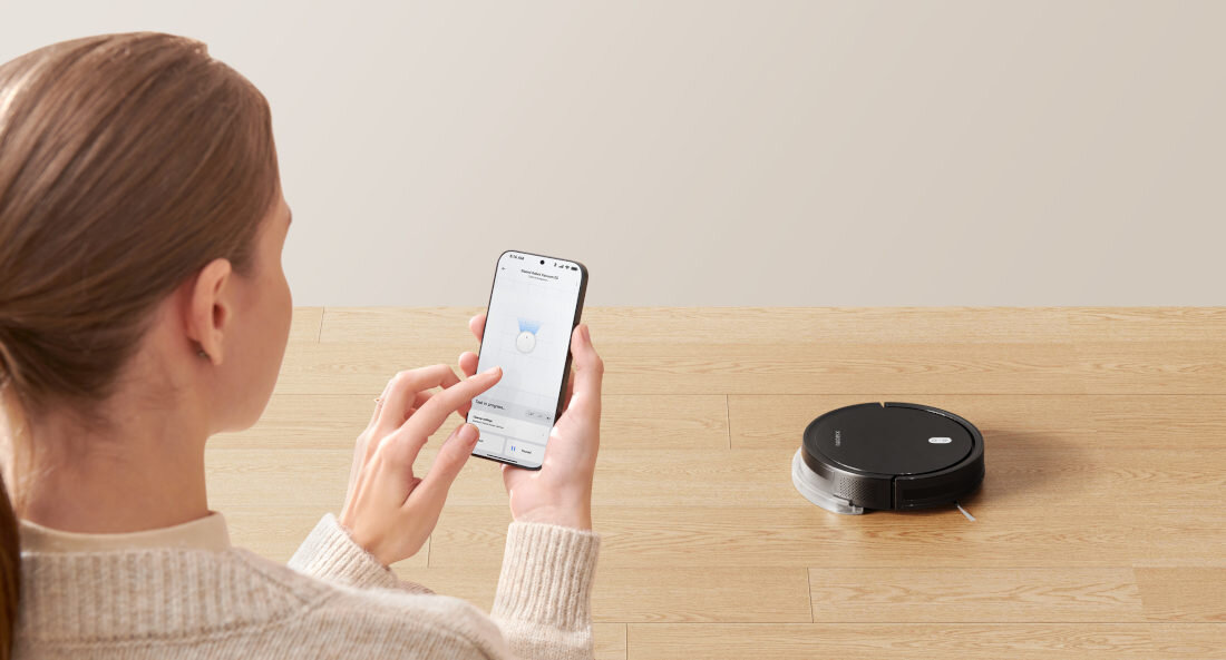 Osoba trzyma smartfon z aplikacją do sterowania robotem sprzątającym. Na podłodze widoczny jest czarny robot sprzątający.